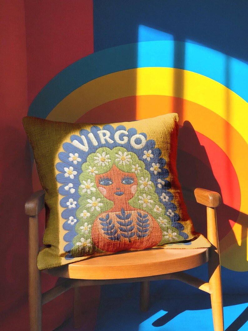 A square throw pillow featuring a vibrant Virgo zodiac design on a golden mustard yellow background. The central illustration shows a terracotta-skinned figure with green hair adorned with white daisies. Blue leaf motifs are printed on the figure’s chest. The word "VIRGO" appears in white bubble letters at the top. The entire design is framed by a royal blue scalloped border, featuring a high-fidelity trompe-l'œil print that mimics 3D embroidery.