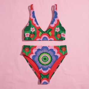 May include: A two-piece swimsuit set with a bold floral pattern. The top has a V-neck design, and the bottoms are high-waisted. The colour palette includes red, green, blue, pink, and white, creating a striking, eye-catching look.