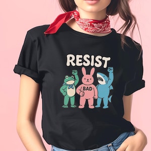 May include: Black t-shirt with the word "RESIST" in white block letters. Below the text are cartoon characters: a green frog, a pink bunny with the word "BAD", and a blue shark, all with raised fists. The shirt has rolled-up sleeves.
