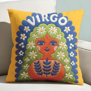 A square throw pillow featuring a vibrant Virgo zodiac design on a golden mustard yellow background. The central illustration shows a terracotta-skinned figure with green hair adorned with white daisies. Blue leaf motifs are printed on the figure’s chest. The word "VIRGO" appears in white bubble letters at the top. The entire design is framed by a royal blue scalloped border, featuring a high-fidelity trompe-l'œil print that mimics 3D embroidery.