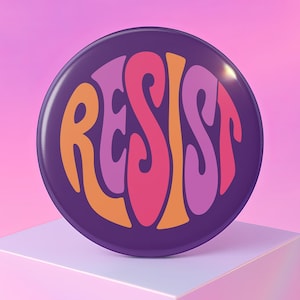 Peut inclure: Un bouton rond violet avec le mot "RESIST" dans une police rétro colorée. Le texte est dans des tons orange, rose et violet. Le bouton est posé sur un fond rose dégradé et repose sur un cube blanc.