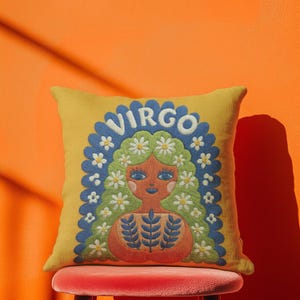 A square throw pillow featuring a vibrant Virgo zodiac design on a golden mustard yellow background. The central illustration shows a terracotta-skinned figure with green hair adorned with white daisies. Blue leaf motifs are printed on the figure’s chest. The word "VIRGO" appears in white bubble letters at the top. The entire design is framed by a royal blue scalloped border, featuring a high-fidelity trompe-l'œil print that mimics 3D embroidery.