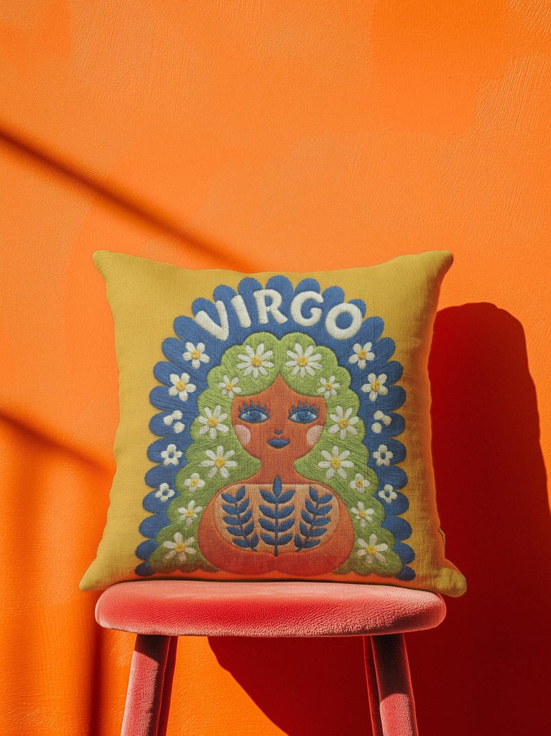 A square throw pillow featuring a vibrant Virgo zodiac design on a golden mustard yellow background. The central illustration shows a terracotta-skinned figure with green hair adorned with white daisies. Blue leaf motifs are printed on the figure’s chest. The word "VIRGO" appears in white bubble letters at the top. The entire design is framed by a royal blue scalloped border, featuring a high-fidelity trompe-l'œil print that mimics 3D embroidery.