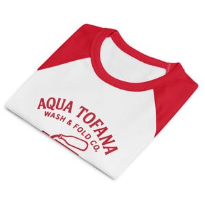 A women's fitted, mid-length micro rib raglan baby tee. The tee's body is solid white, made from a 1x1 micro-rib fabric.  It features short, set-in sleeves and a crewneck collar binding in a bold, contrasting athletic red. Centered on the chest is a vintage-inspired graphic printed in the same red ink. The design is for the "Aqua Tofana Wash & Fold Co." and features an illustration of an old-fashioned washing machine. Text below the machine reads "Making small problems go away" and "Since 1680."
