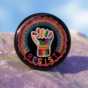 May include: A black button with a crocheted rainbow fist and the word "RESIST" in white.