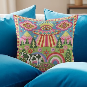 Description of a square throw pillow with a vibrant maximalist alien design. A central UFO with a large all-seeing eye hovers above a landscape with two rainbows, pink mountains, and green pine trees. A black and white spotted cow stands in the foreground. The design is a high-contrast palette of sky blue, magenta, hot pink, and meadow green. The pillow has a faux-embroidery 2D print finish on linen-feel fabric, framed by a yellow and pink scalloped floral border.