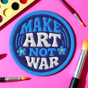 May include: Circular embroidered patch with the slogan "MAKE ART NOT WAR" in white and blue lettering. The patch has a blue border and a dark purple centre, with small blue flowers. Paintbrushes and a paint palette are arranged around the patch on a pink surface.