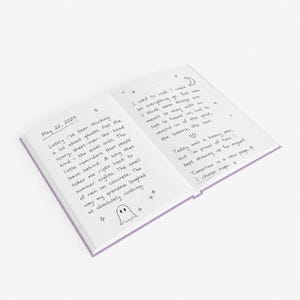 A 5 by 7 inch hardcover journal with a matte pastel purple cover and black printed graphics. The front features a retro inspired illustration of a white ghost character. One design shows the ghost holding a locked book flanked by a lit candle and a skeleton key with text reading Notes For My Future Ghost. The other shows the ghost peeking over a blank label with text reading Property Of The Future Ghost. Both include crescent moons, four point stars, and vintage decorative border frames.