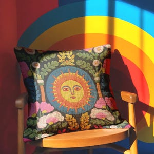 A square folk art sun throw pillow with a dark, charcoal-gray background. The central design is a vibrant celestial sun face in shades of orange, yellow, and red, with defined facial features. The sun is enclosed in a circular blue border with a textured, faux-embroidery pattern. Surrounding the central sun are symmetrical, colorful botanical and floral elements, including pink and white flowers, and green and gold leaves. The overall style is maximalist and bohemian.