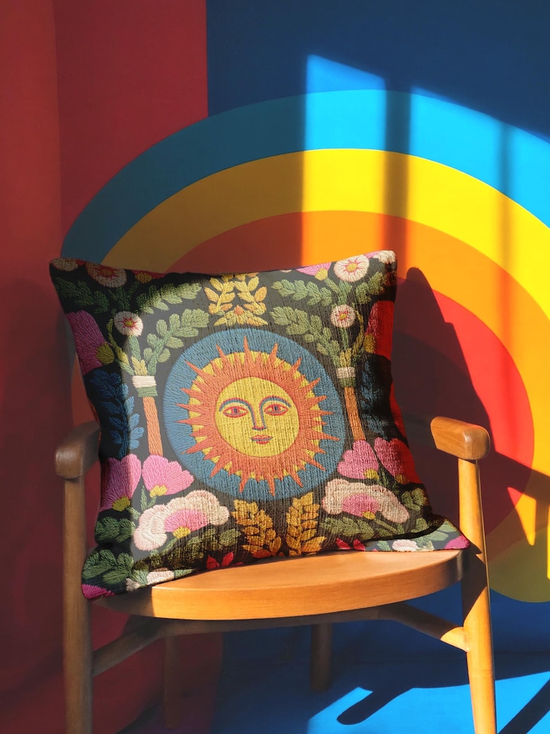 A square folk art sun throw pillow with a dark, charcoal-gray background. The central design is a vibrant celestial sun face in shades of orange, yellow, and red, with defined facial features. The sun is enclosed in a circular blue border with a textured, faux-embroidery pattern. Surrounding the central sun are symmetrical, colorful botanical and floral elements, including pink and white flowers, and green and gold leaves. The overall style is maximalist and bohemian.
