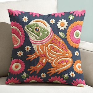 May include: A square throw pillow with a dark blue fabric base featuring an embroidered design of a green and orange frog wearing a white astronaut helmet. The frog is surrounded by colorful embroidered flowers in shades of pink, orange, and white.