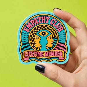 May include: A sticker with a light blue background featuring the words "EMPATHY CLUB" and "LIFETIME MEMBER" in a retro font. The design includes two yellow profile faces, an eye, and a checkered pattern.