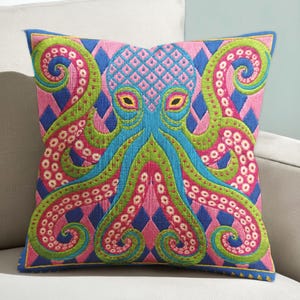 A square decorative throw pillow cover featuring a highly symmetrical bright octopus design. The octopus has a turquoise blue body with a diamond patterned head and eight curled tentacles detailed in bright pink and lime green. The background is a repeating diamond argyle pattern in royal blue and light pink. The artwork is a flat two dimensional print that mimics the textured look of intricate crewelwork embroidery. The pillow features a hidden zipper on the bottom edge. No insert included.