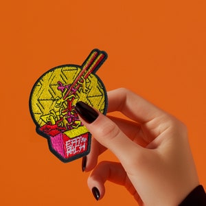 Könnte beinhalten: Ein gelber und roter gestickter Aufnäher mit einem Cartoon-Bild eines Take-Away-Behälters gefüllt mit Nudeln und Essstäbchen. Der Text "Eat the Rich" ist auf den Behälter gestickt.