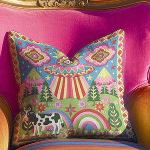 Description of a square throw pillow with a vibrant maximalist alien design. A central UFO with a large all-seeing eye hovers above a landscape with two rainbows, pink mountains, and green pine trees. A black and white spotted cow stands in the foreground. The design is a high-contrast palette of sky blue, magenta, hot pink, and meadow green. The pillow has a faux-embroidery 2D print finish on linen-feel fabric, framed by a yellow and pink scalloped floral border.