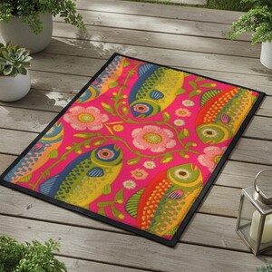 Rectangular indoor outdoor doormat featuring a vibrant hot pink background with a symmetrical maximalist pattern. The design showcases stylized sardines in yellow, lime green, and cobalt blue swimming horizontally towards a central vertical axis of blooming pale pink flowers and green vines. The imagery mimics the texture of vintage needlepoint embroidery but is a flat print on polyester. The mat is framed by a sleek black trim and features a non-slip rubber backing for safety.