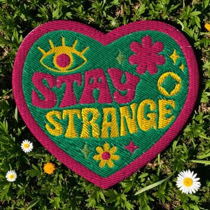 May include: Embroidered heart-shaped patch with the words "STAY STRANGE" in yellow and pink. The patch features a green background with a pink border, and includes embroidered flowers, stars, and an eye design. The patch is set against a background of green grass.