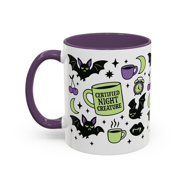 White ceramic mug with vibrant purple interior and matching C-handle. Features a full wrap-around, 'spooky cute' pastel goth design. Central graphic is a lime green coffee cup with 'CERTIFIED NIGHT CREATURE' in black text. Surrounding illustrations include grumpy black bats, a steaming green coffee cup, a purple mug, vampire fangs, crescent moon, black cherries, and an alarm clock. Design uses bold graphics in black, lime green, and purple, accented with small black stars. Pastel Goth vibes.