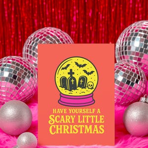 A vibrant, flat-lay photograph of the "Scary Little Christmas" greeting card. The card has a bright orange background with bold, retro yellow text. The central graphic is a snow globe with a yellow glass dome and a hot pink base. Inside the globe is a black silhouette of a spooky graveyard scene, featuring tombstones with crosses, a skull, and a swarm of flying bats. The card is made from premium 120lb silk cover stock with a slight sheen and measures 5x7 inches.