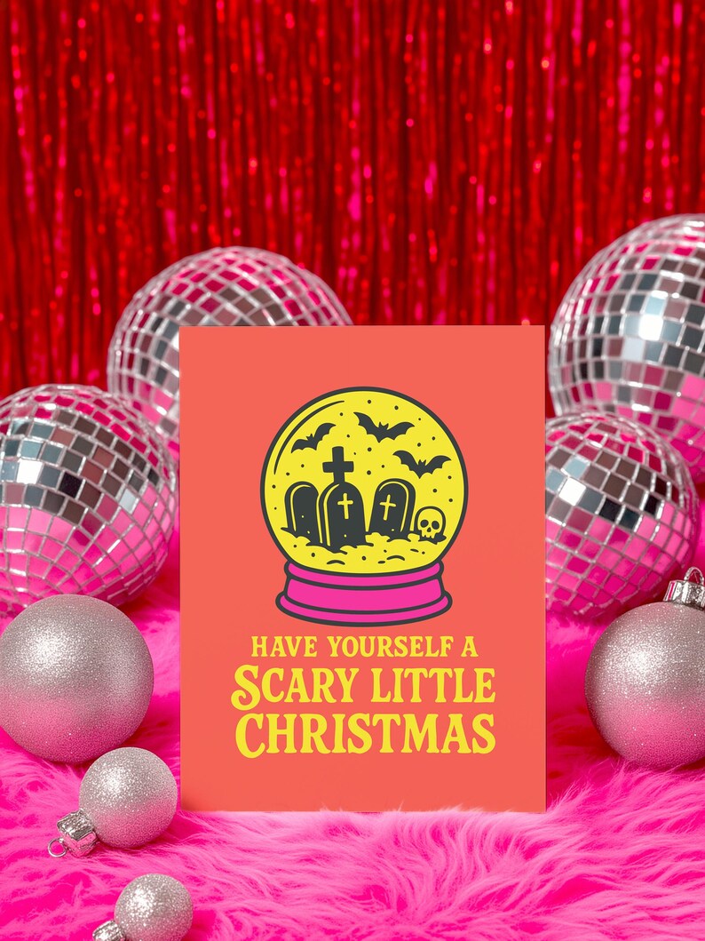 A vibrant, flat-lay photograph of the "Scary Little Christmas" greeting card. The card has a bright orange background with bold, retro yellow text. The central graphic is a snow globe with a yellow glass dome and a hot pink base. Inside the globe is a black silhouette of a spooky graveyard scene, featuring tombstones with crosses, a skull, and a swarm of flying bats. The card is made from premium 120lb silk cover stock with a slight sheen and measures 5x7 inches.