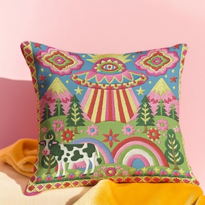 Description of a square throw pillow with a vibrant maximalist alien design. A central UFO with a large all-seeing eye hovers above a landscape with two rainbows, pink mountains, and green pine trees. A black and white spotted cow stands in the foreground. The design is a high-contrast palette of sky blue, magenta, hot pink, and meadow green. The pillow has a faux-embroidery 2D print finish on linen-feel fabric, framed by a yellow and pink scalloped floral border.