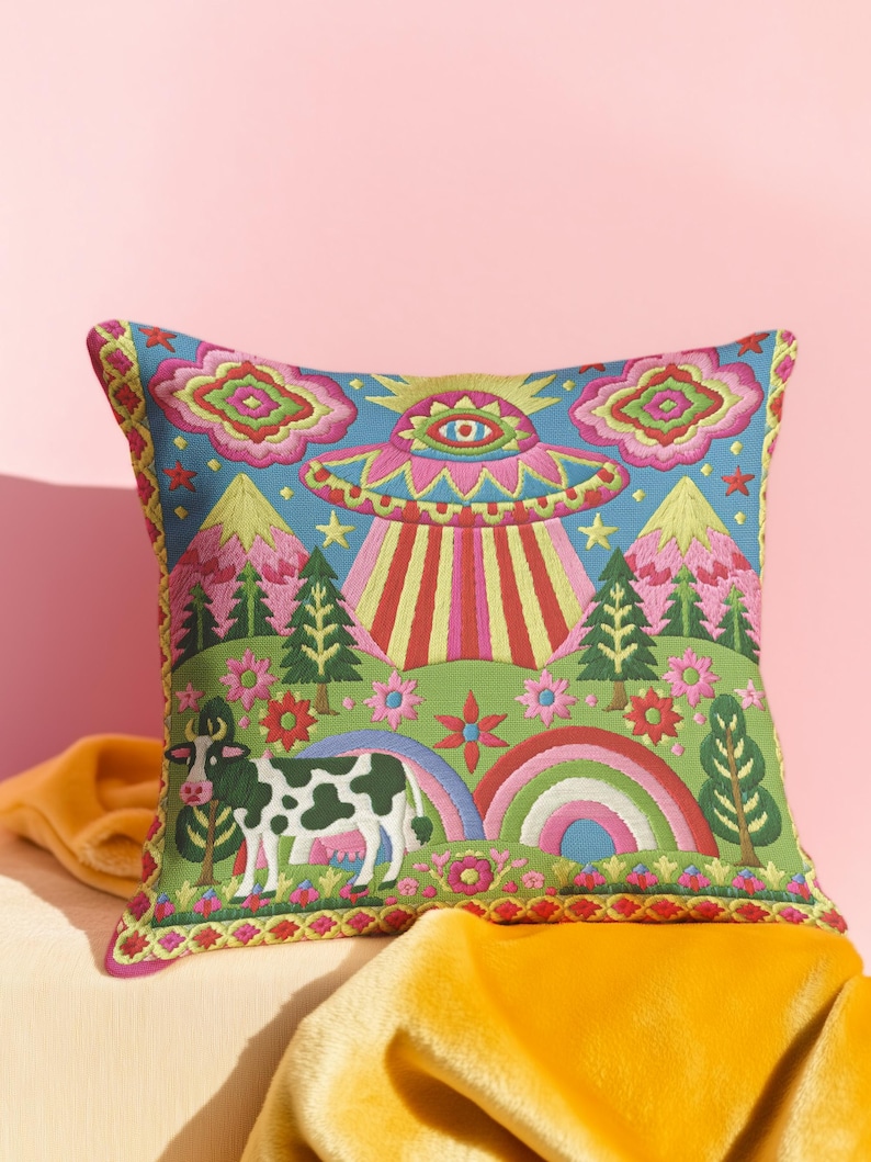 Description of a square throw pillow with a vibrant maximalist alien design. A central UFO with a large all-seeing eye hovers above a landscape with two rainbows, pink mountains, and green pine trees. A black and white spotted cow stands in the foreground. The design is a high-contrast palette of sky blue, magenta, hot pink, and meadow green. The pillow has a faux-embroidery 2D print finish on linen-feel fabric, framed by a yellow and pink scalloped floral border.