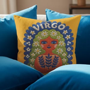 A square throw pillow featuring a vibrant Virgo zodiac design on a golden mustard yellow background. The central illustration shows a terracotta-skinned figure with green hair adorned with white daisies. Blue leaf motifs are printed on the figure’s chest. The word "VIRGO" appears in white bubble letters at the top. The entire design is framed by a royal blue scalloped border, featuring a high-fidelity trompe-l'œil print that mimics 3D embroidery.