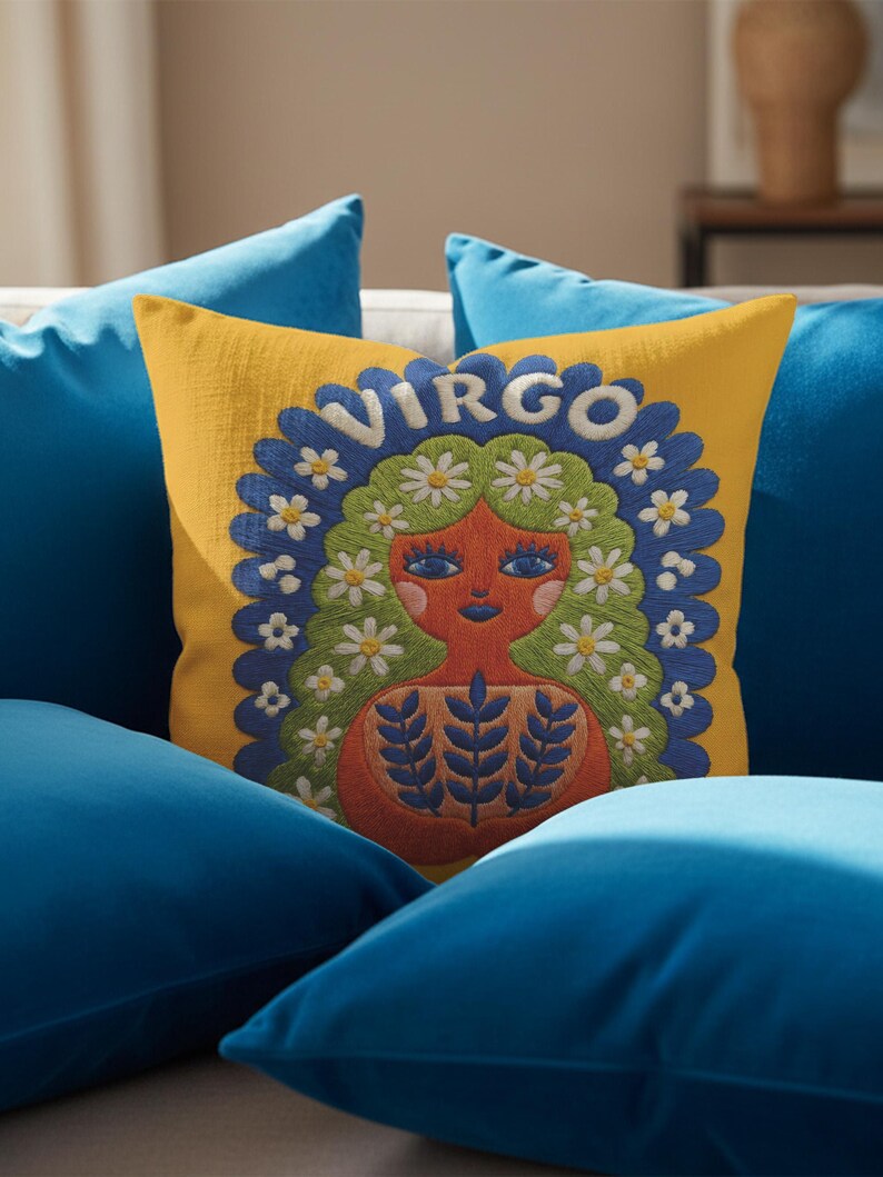 A square throw pillow featuring a vibrant Virgo zodiac design on a golden mustard yellow background. The central illustration shows a terracotta-skinned figure with green hair adorned with white daisies. Blue leaf motifs are printed on the figure’s chest. The word "VIRGO" appears in white bubble letters at the top. The entire design is framed by a royal blue scalloped border, featuring a high-fidelity trompe-l'œil print that mimics 3D embroidery.