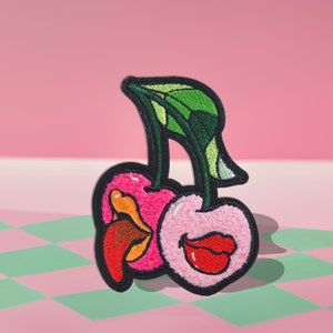 May include: Two embroidered cherries with pink and red lips, one with a brown tongue sticking out, on a green and pink checkered background.