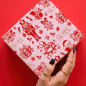 Premium satin finish wrapping paper featuring a repeating pastel goth pattern on a pink background. The design showcases a playful yet spooky aesthetic with high-contrast skeleton nutcrackers, skull jack-in-the-boxes, and rocking horse skeletons in soft pinks, and reds. The paper texture appears semi-glossy. Shown in three available sizes including sheets and rolls, specifically 29 inches wide, suitable for wrapping gifts for holidays, birthdays, or gothic themed occasions.
