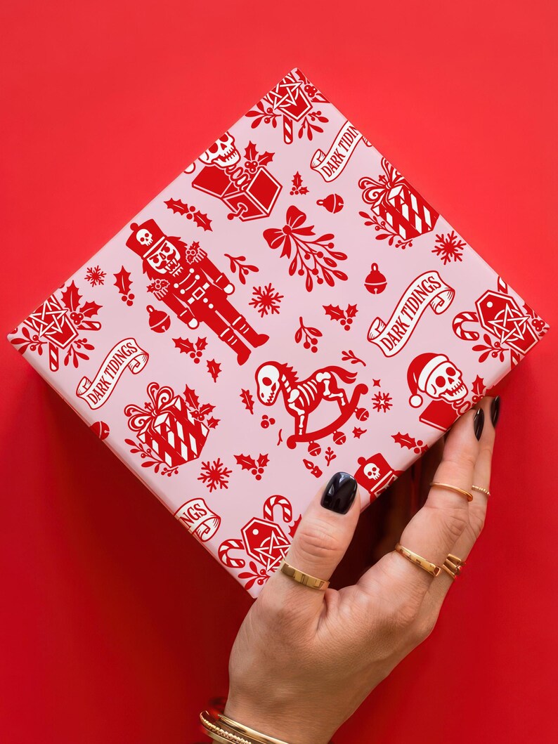 Premium satin finish wrapping paper featuring a repeating pastel goth pattern on a pink background. The design showcases a playful yet spooky aesthetic with high-contrast skeleton nutcrackers, skull jack-in-the-boxes, and rocking horse skeletons in soft pinks, and reds. The paper texture appears semi-glossy. Shown in three available sizes including sheets and rolls, specifically 29 inches wide, suitable for wrapping gifts for holidays, birthdays, or gothic themed occasions.