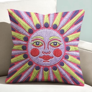 Peut inclure: Un coussin carré orné d'un motif de visage de soleil brodé sur un fond rose. Le soleil a un visage violet avec des joues et des lèvres rouges, entouré de rayons alternés vert clair et violets avec des accents circulaires violet foncé.