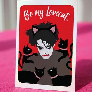 May include: A greeting card with a red background and the text "Be my Lovecat." The card features a stylized illustration of a person with black hair and makeup, wearing cat ears, surrounded by black cats with pink collars.