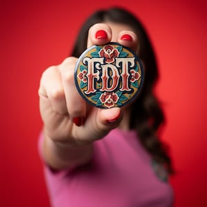 May include: A round button with the letters "FDT" in a decorative font, surrounded by floral designs. The button has a navy blue border and red, gold, and teal accents. The background is a solid red.