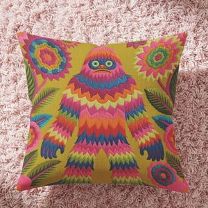 A square faux embroidery pillow featuring a brightly colored maximalist design against a mustard yellow background. In the center stands a stylized, furry Bigfoot or Sasquatch figure rendered in vibrant layers of pink, orange, blue, and lime green with a blue face and yellow eyes. The central creature is surrounded by large, detailed symmetrical pink and blue flowers and leaves. The flat printed fabric creates a highly realistic optical illusion of dimensional crewel embroidery stitches.