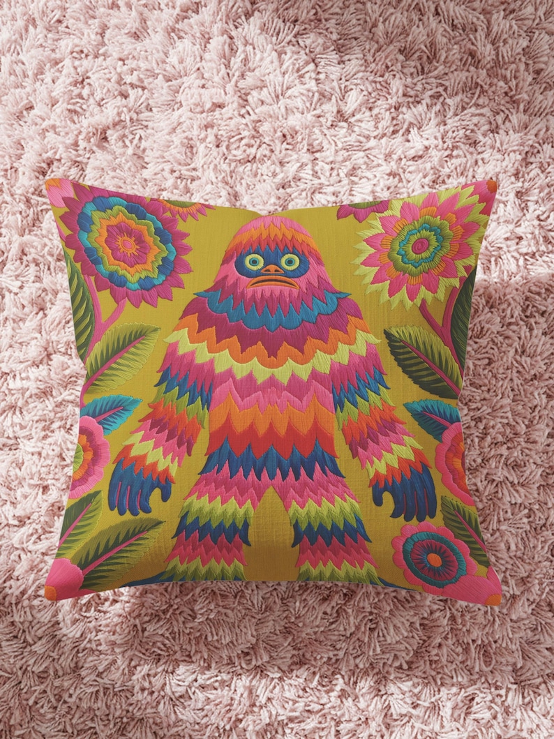 A square faux embroidery pillow featuring a brightly colored maximalist design against a mustard yellow background. In the center stands a stylized, furry Bigfoot or Sasquatch figure rendered in vibrant layers of pink, orange, blue, and lime green with a blue face and yellow eyes. The central creature is surrounded by large, detailed symmetrical pink and blue flowers and leaves. The flat printed fabric creates a highly realistic optical illusion of dimensional crewel embroidery stitches.