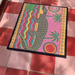 A rectangular doormat featuring a colorful 2D flat print designed to mimic raised, textured yarn embroidery. The central graphic showcases a large dark grey and yellow alligator resting in wavy water stripes of red, bright blue, and yellow. The background is hot pink, decorated with two green palm trees, a leafy green agave plant, and two stylized suns. The entire design is enclosed by a yellow border with repeating circle motifs and finished with a sleek black outer edge border trim.