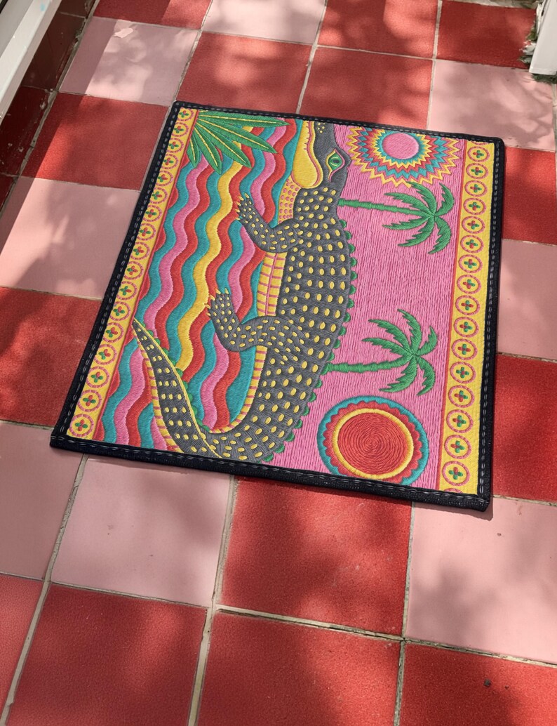 A rectangular doormat featuring a colorful 2D flat print designed to mimic raised, textured yarn embroidery. The central graphic showcases a large dark grey and yellow alligator resting in wavy water stripes of red, bright blue, and yellow. The background is hot pink, decorated with two green palm trees, a leafy green agave plant, and two stylized suns. The entire design is enclosed by a yellow border with repeating circle motifs and finished with a sleek black outer edge border trim.