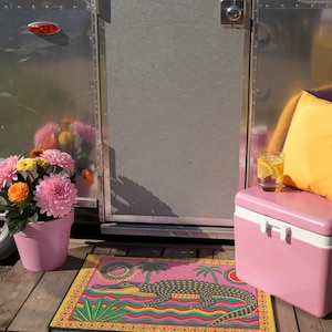 A rectangular doormat featuring a colorful 2D flat print designed to mimic raised, textured yarn embroidery. The central graphic showcases a large dark grey and yellow alligator resting in wavy water stripes of red, bright blue, and yellow. The background is hot pink, decorated with two green palm trees, a leafy green agave plant, and two stylized suns. The entire design is enclosed by a yellow border with repeating circle motifs and finished with a sleek black outer edge border trim.