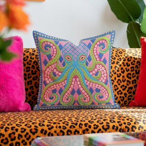 A square decorative throw pillow cover featuring a highly symmetrical bright octopus design. The octopus has a turquoise blue body with a diamond patterned head and eight curled tentacles detailed in bright pink and lime green. The background is a repeating diamond argyle pattern in royal blue and light pink. The artwork is a flat two dimensional print that mimics the textured look of intricate crewelwork embroidery. The pillow features a hidden zipper on the bottom edge. No insert included.