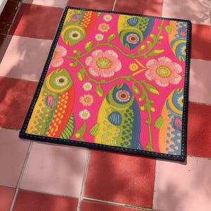 Rectangular indoor outdoor doormat featuring a vibrant hot pink background with a symmetrical maximalist pattern. The design showcases stylized sardines in yellow, lime green, and cobalt blue swimming horizontally towards a central vertical axis of blooming pale pink flowers and green vines. The imagery mimics the texture of vintage needlepoint embroidery but is a flat print on polyester. The mat is framed by a sleek black trim and features a non-slip rubber backing for safety.