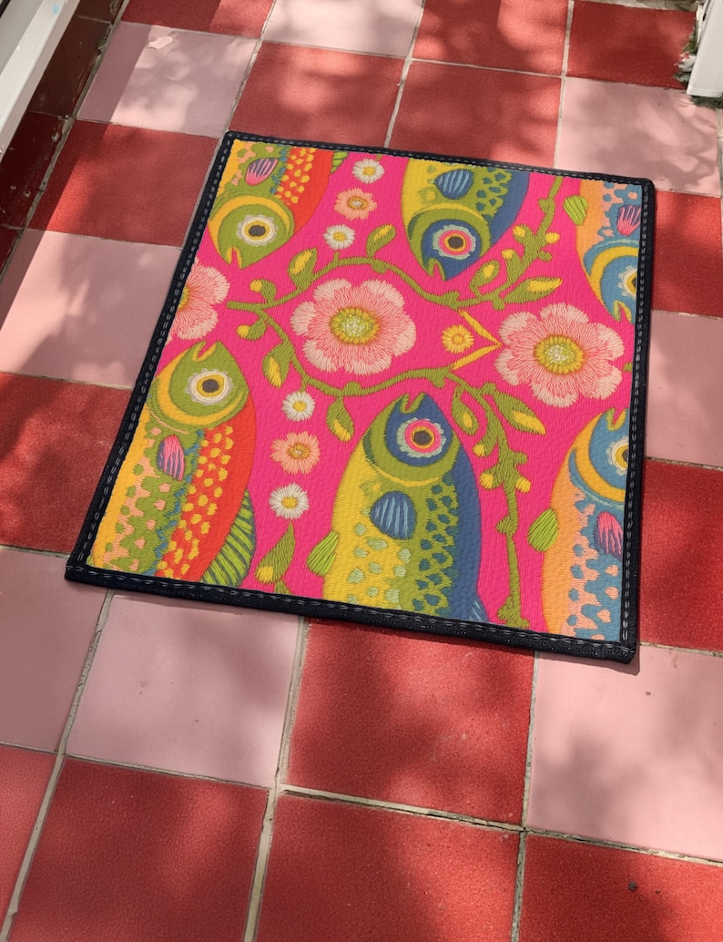 Rectangular indoor outdoor doormat featuring a vibrant hot pink background with a symmetrical maximalist pattern. The design showcases stylized sardines in yellow, lime green, and cobalt blue swimming horizontally towards a central vertical axis of blooming pale pink flowers and green vines. The imagery mimics the texture of vintage needlepoint embroidery but is a flat print on polyester. The mat is framed by a sleek black trim and features a non-slip rubber backing for safety.
