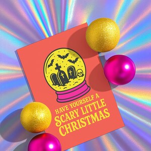 A vibrant, flat-lay photograph of the "Scary Little Christmas" greeting card. The card has a bright orange background with bold, retro yellow text. The central graphic is a snow globe with a yellow glass dome and a hot pink base. Inside the globe is a black silhouette of a spooky graveyard scene, featuring tombstones with crosses, a skull, and a swarm of flying bats. The card is made from premium 120lb silk cover stock with a slight sheen and measures 5x7 inches.