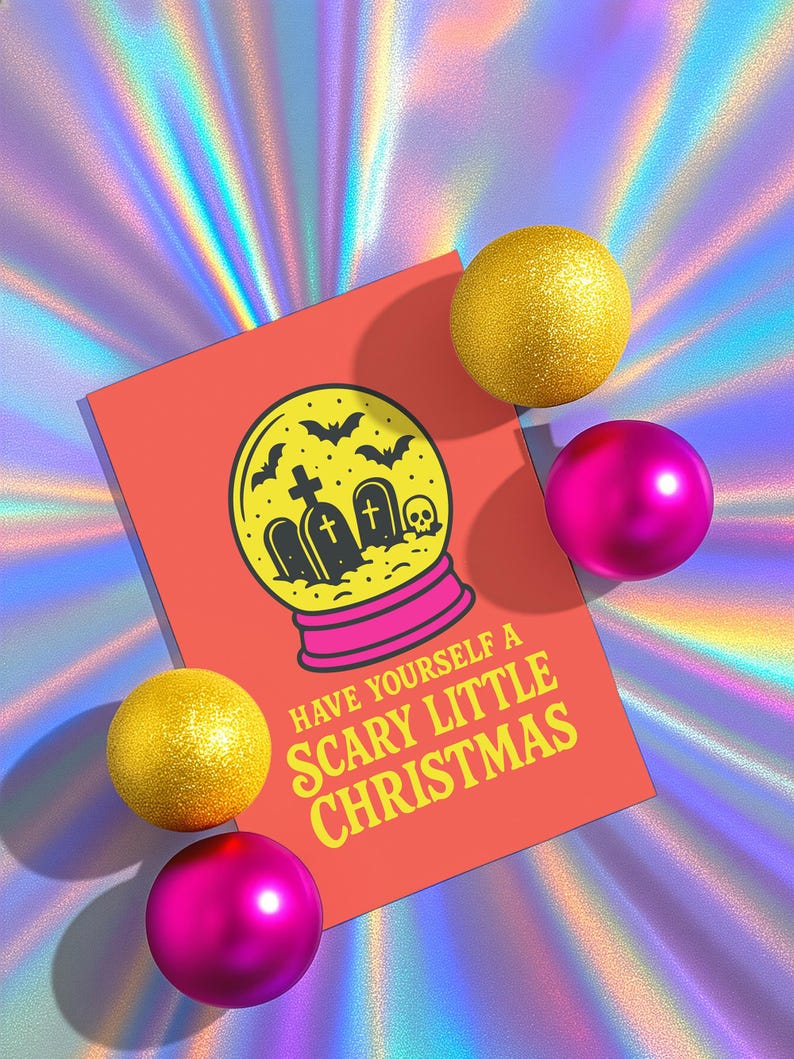 A vibrant, flat-lay photograph of the "Scary Little Christmas" greeting card. The card has a bright orange background with bold, retro yellow text. The central graphic is a snow globe with a yellow glass dome and a hot pink base. Inside the globe is a black silhouette of a spooky graveyard scene, featuring tombstones with crosses, a skull, and a swarm of flying bats. The card is made from premium 120lb silk cover stock with a slight sheen and measures 5x7 inches.