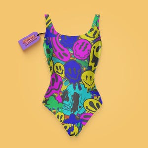May include: A one-piece swimsuit featuring a vibrant, multi-colored graphic print of melting smiley faces and skulls in shades of yellow, pink, blue, and green. A purple rectangular tag attached to the strap displays the text Honey Dazed.