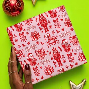 Premium satin finish wrapping paper featuring a repeating pastel goth pattern on a pink background. The design showcases a playful yet spooky aesthetic with high-contrast skeleton nutcrackers, skull jack-in-the-boxes, and rocking horse skeletons in soft pinks, and reds. The paper texture appears semi-glossy. Shown in three available sizes including sheets and rolls, specifically 29 inches wide, suitable for wrapping gifts for holidays, birthdays, or gothic themed occasions.