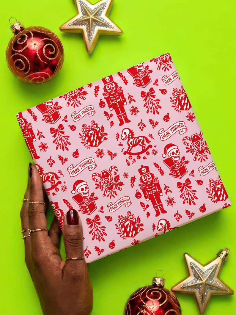 Premium satin finish wrapping paper featuring a repeating pastel goth pattern on a pink background. The design showcases a playful yet spooky aesthetic with high-contrast skeleton nutcrackers, skull jack-in-the-boxes, and rocking horse skeletons in soft pinks, and reds. The paper texture appears semi-glossy. Shown in three available sizes including sheets and rolls, specifically 29 inches wide, suitable for wrapping gifts for holidays, birthdays, or gothic themed occasions.
