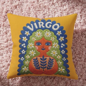 A square throw pillow featuring a vibrant Virgo zodiac design on a golden mustard yellow background. The central illustration shows a terracotta-skinned figure with green hair adorned with white daisies. Blue leaf motifs are printed on the figure’s chest. The word "VIRGO" appears in white bubble letters at the top. The entire design is framed by a royal blue scalloped border, featuring a high-fidelity trompe-l'œil print that mimics 3D embroidery.