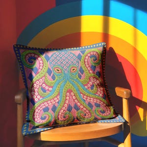 A square decorative throw pillow cover featuring a highly symmetrical bright octopus design. The octopus has a turquoise blue body with a diamond patterned head and eight curled tentacles detailed in bright pink and lime green. The background is a repeating diamond argyle pattern in royal blue and light pink. The artwork is a flat two dimensional print that mimics the textured look of intricate crewelwork embroidery. The pillow features a hidden zipper on the bottom edge. No insert included.