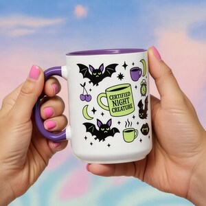 White ceramic mug with vibrant purple interior and matching C-handle. Features a full wrap-around, 'spooky cute' pastel goth design. Central graphic is a lime green coffee cup with 'CERTIFIED NIGHT CREATURE' in black text. Surrounding illustrations include grumpy black bats, a steaming green coffee cup, a purple mug, vampire fangs, crescent moon, black cherries, and an alarm clock. Design uses bold graphics in black, lime green, and purple, accented with small black stars. Pastel Goth vibes.